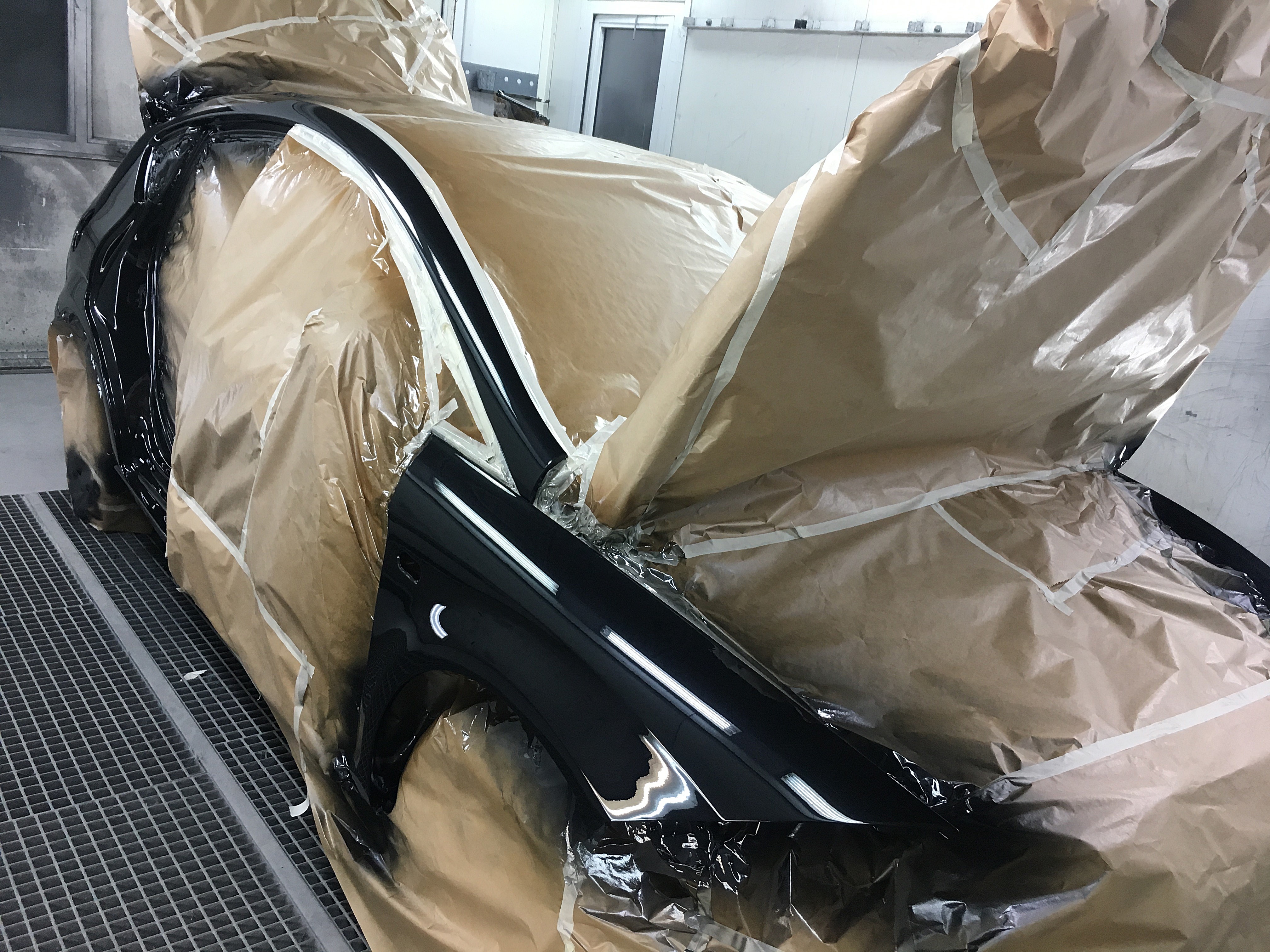 Elementy karoserii samochodowej, błotnik i drzwi, po lakierowaniu na czarno, zabezpieczone papierem i folią malarską w kabinie lakierniczej.