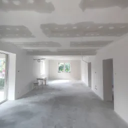 Wnętrze w trakcie prac wykończeniowych: sufit z widocznymi fragmentami płyt, świeżo położona wylewka na podłodze, ściany przygotowane do malowania, widok na okno z zielenią na zewnątrz.