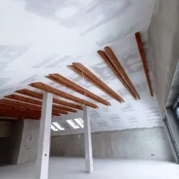 Poddasze w trakcie ocieplania z widocznymi drewnianymi belkami, częściowo pokryte płytami kartonowo-gipsowymi i wstępnie zagruntowane, z oknami dachowymi.