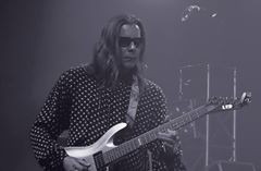 Gitarzysta w okularach przeciwsłonecznych gra na elektrycznej gitarze ESP LTD na scenie oświetlonej punktowym światłem. Mężczyzna ma długie włosy i koszulę w kropki.