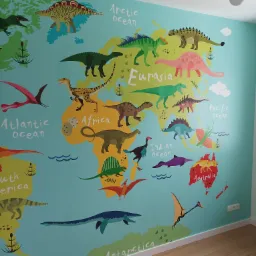 Ściana w pokoju dziecięcym pokryta kolorową tapetą z motywem mapy świata i dinozaurów, widoczne gniazdka elektryczne i drewniana podłoga.