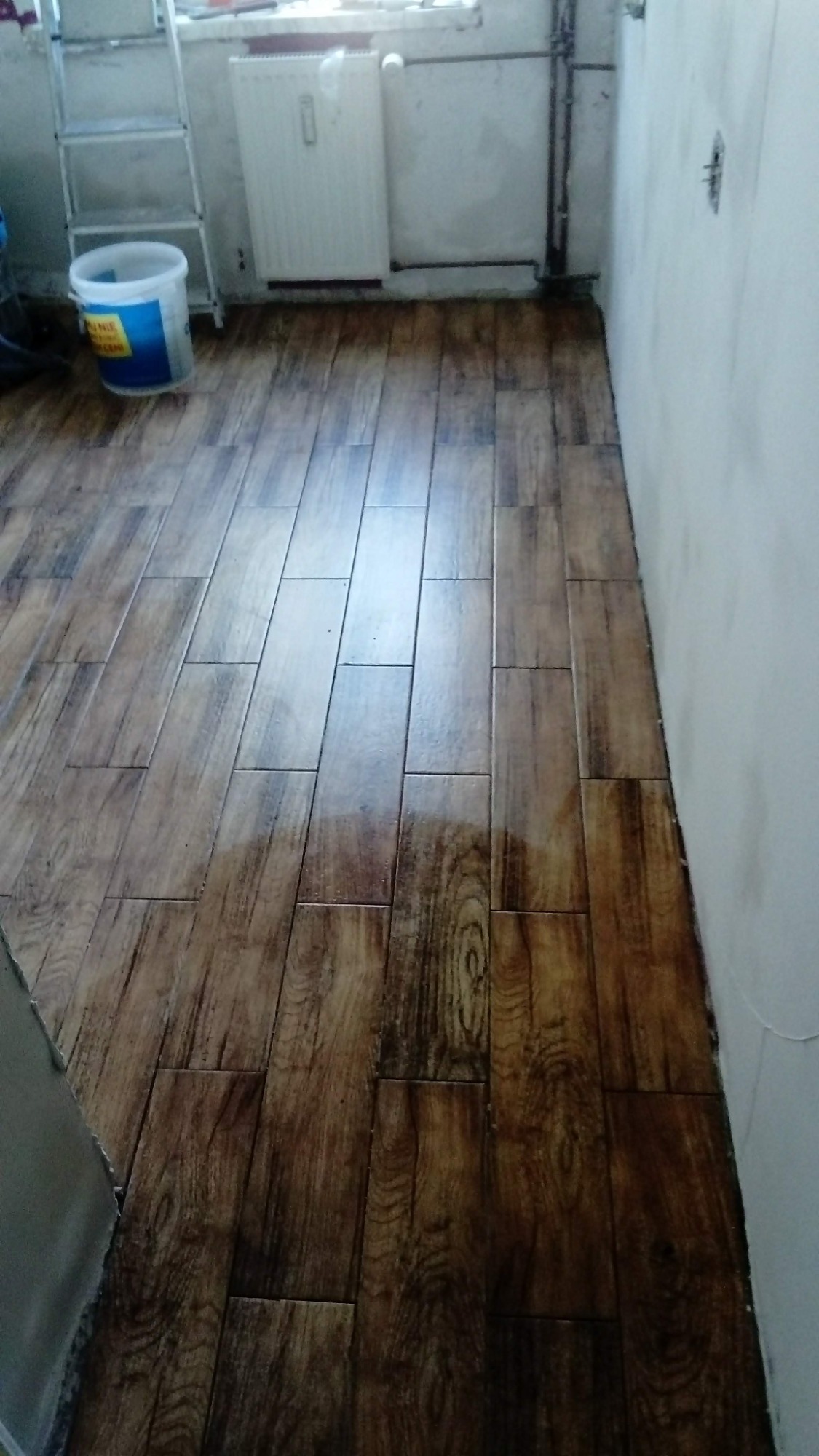 Ułożona podłoga z płytek imitujących drewno w trakcie prac wykończeniowych, widoczne wiadro farby i drabina.