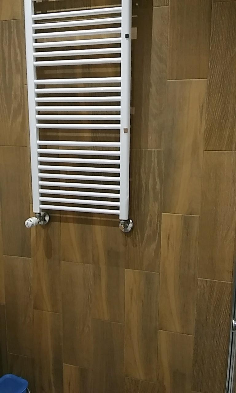 Biały grzejnik drabinkowy zamontowany na ścianie wyłożonej brązowymi płytkami imitującymi drewno w łazience.