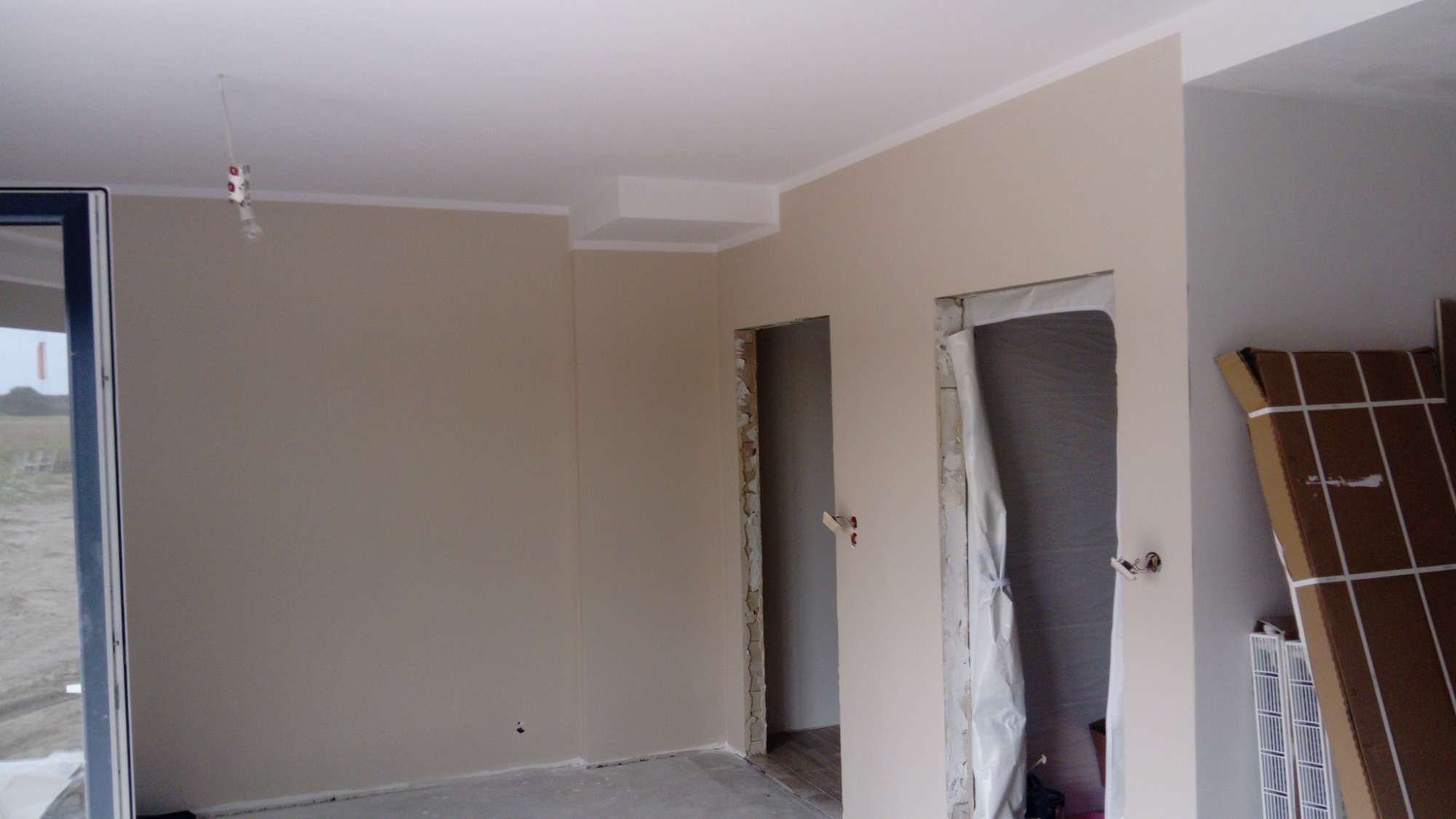 Wnętrze w trakcie prac wykończeniowych: świeżo pomalowane ściany w odcieniach beżu, widoczne surowe ościeżnice drzwiowe zabezpieczone folią, na suficie widoczna prowizoryczna instalacja...