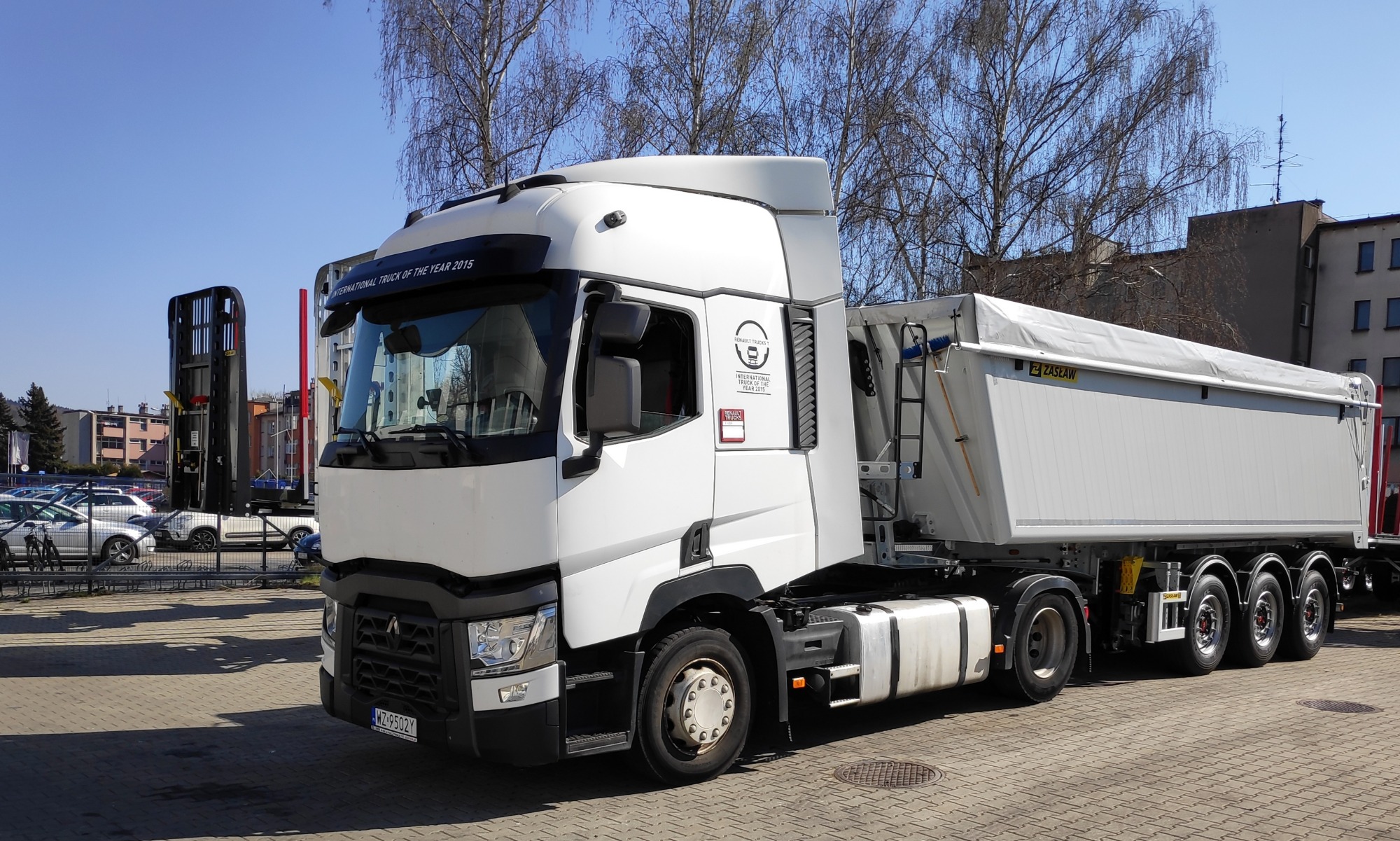 Biały ciągnik siodłowy Renault z naczepą wywrotką ZASŁAW, zaparkowany na placu z kostki brukowej, widoczny napis 'International Truck of the Year 2015' na kabinie.