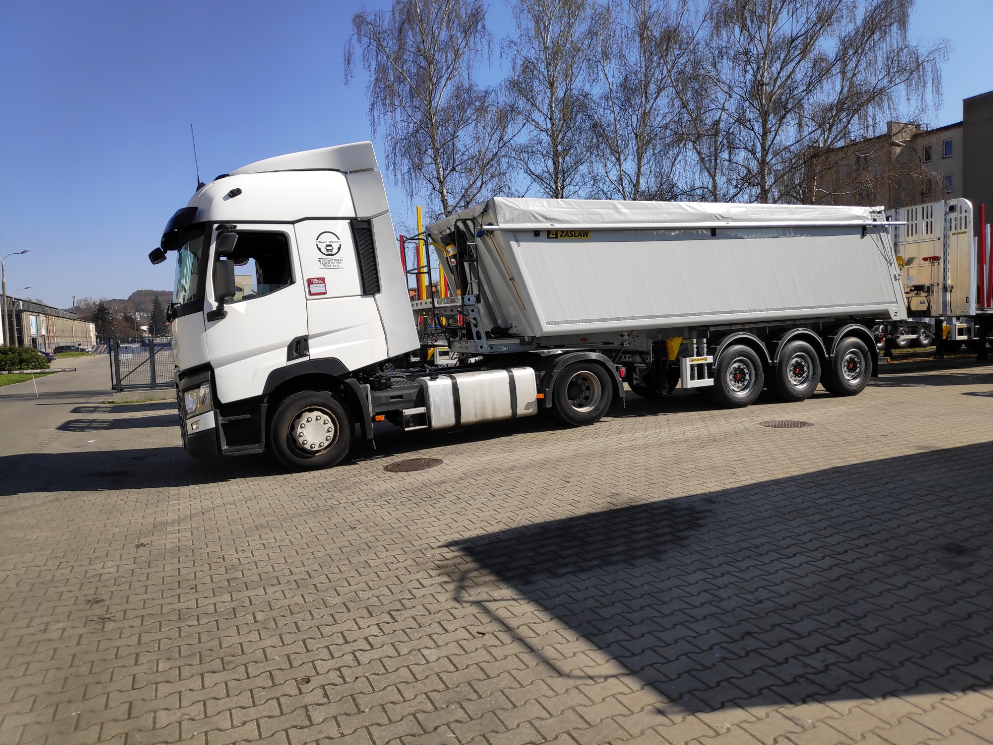 Biała ciężarówka marki Renault z naczepą ZASŁAW stoi na brukowanym placu w słoneczny dzień, widoczne logo producenta na kabinie i elementy konstrukcyjne naczepy.
