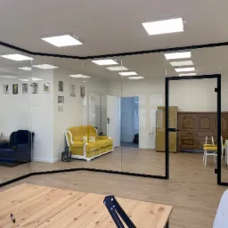 Nowoczesne biuro z przeszklonymi ścianami z czarnymi ramami, widoczne meble w stylu retro, drewniana podłoga i jasne oświetlenie.