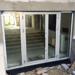 Trzyskrzydłowe drzwi balkonowe z białymi ramami i szklanym wypełnieniem, widoczne od strony placu budowy, z gruzem i cegłami przed progiem.