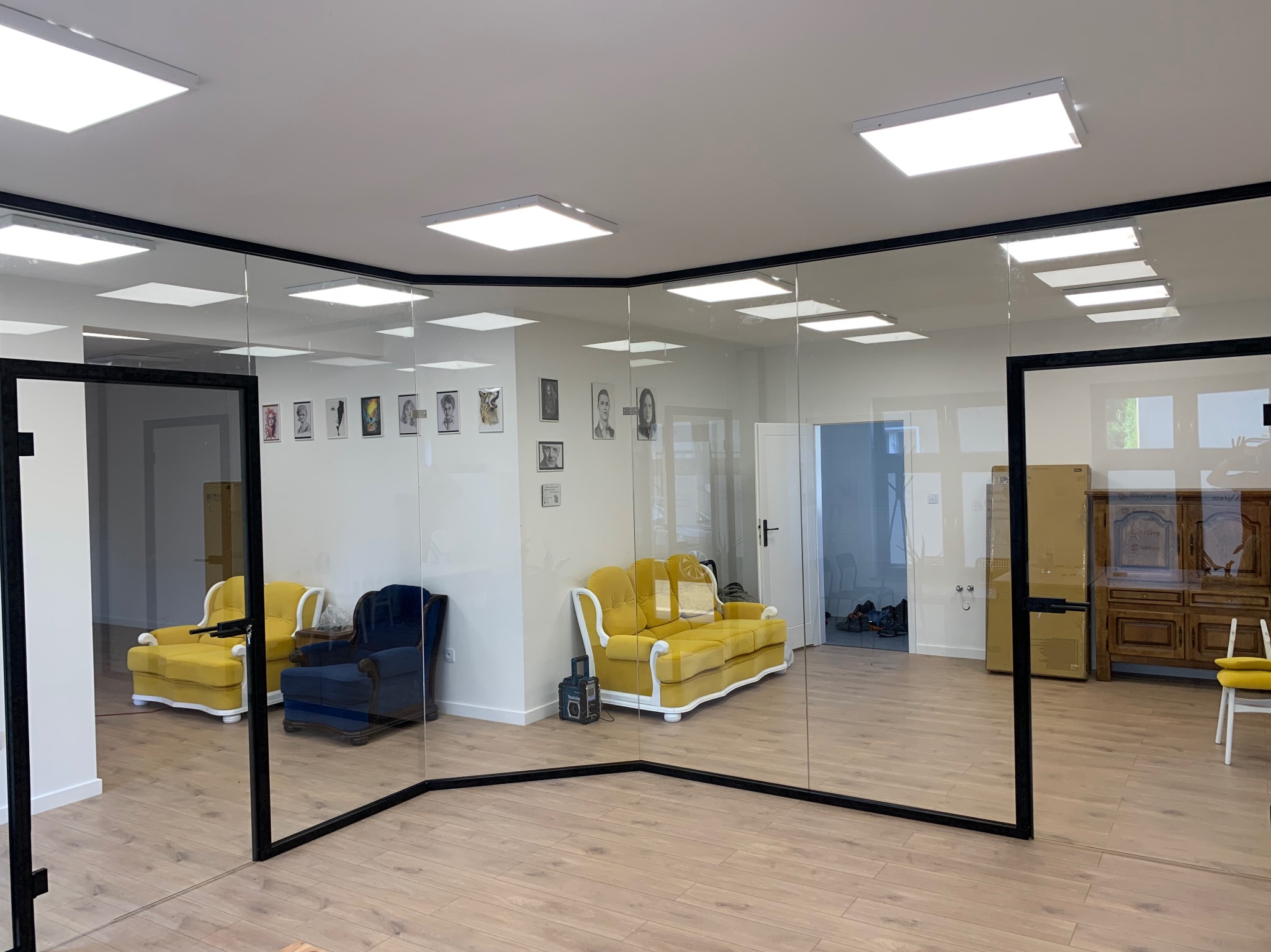 Przeszklona przestrzeń biurowa z czarnymi ramami, widoczne żółte i niebieskie sofy, drewniana podłoga i oświetlenie sufitowe.