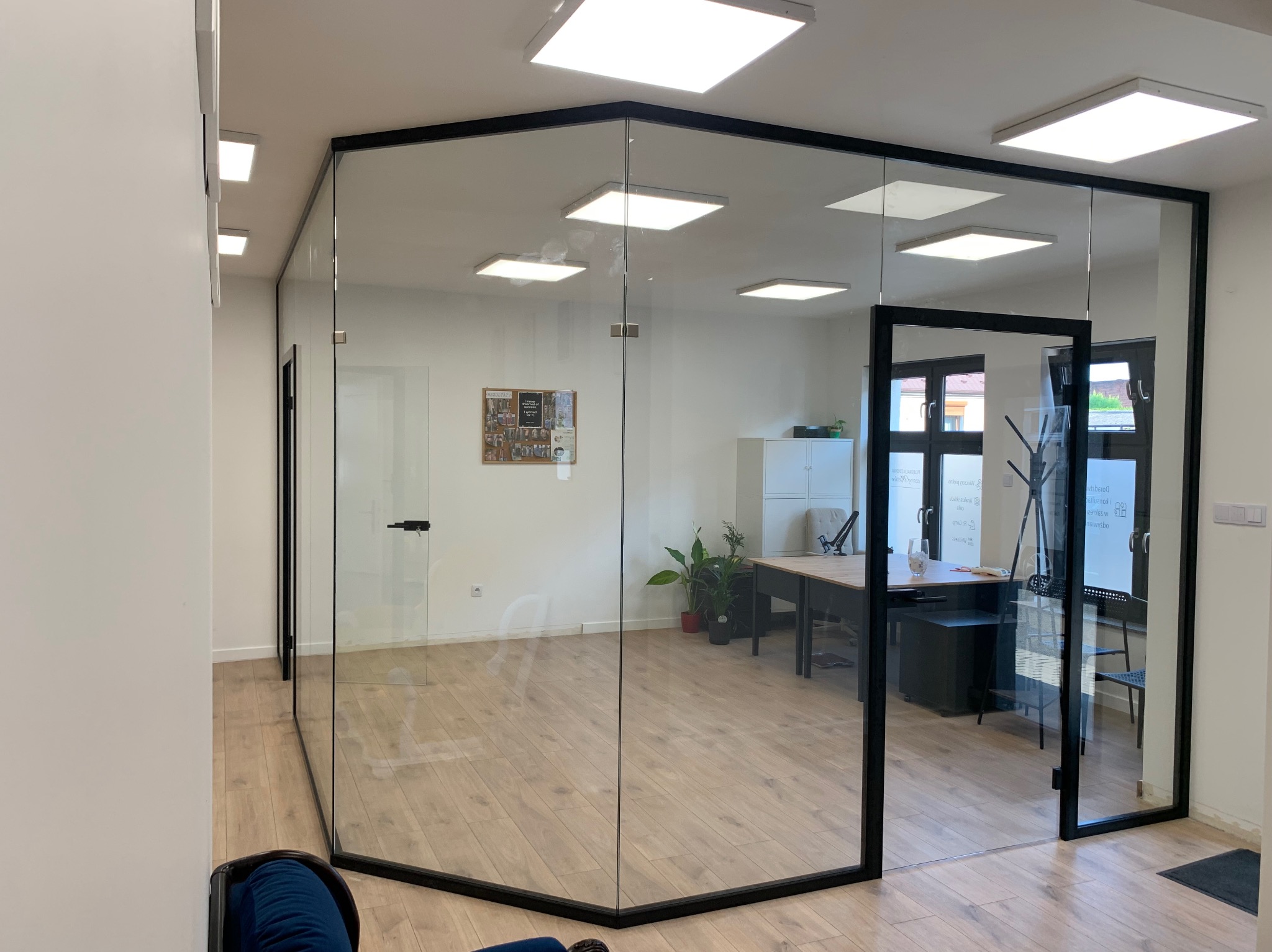 Szklana ścianka działowa z czarną ramą tworząca nowoczesne biuro, widok z zewnątrz na wnętrze z biurkiem i roślinami, jasne oświetlenie sufitowe.