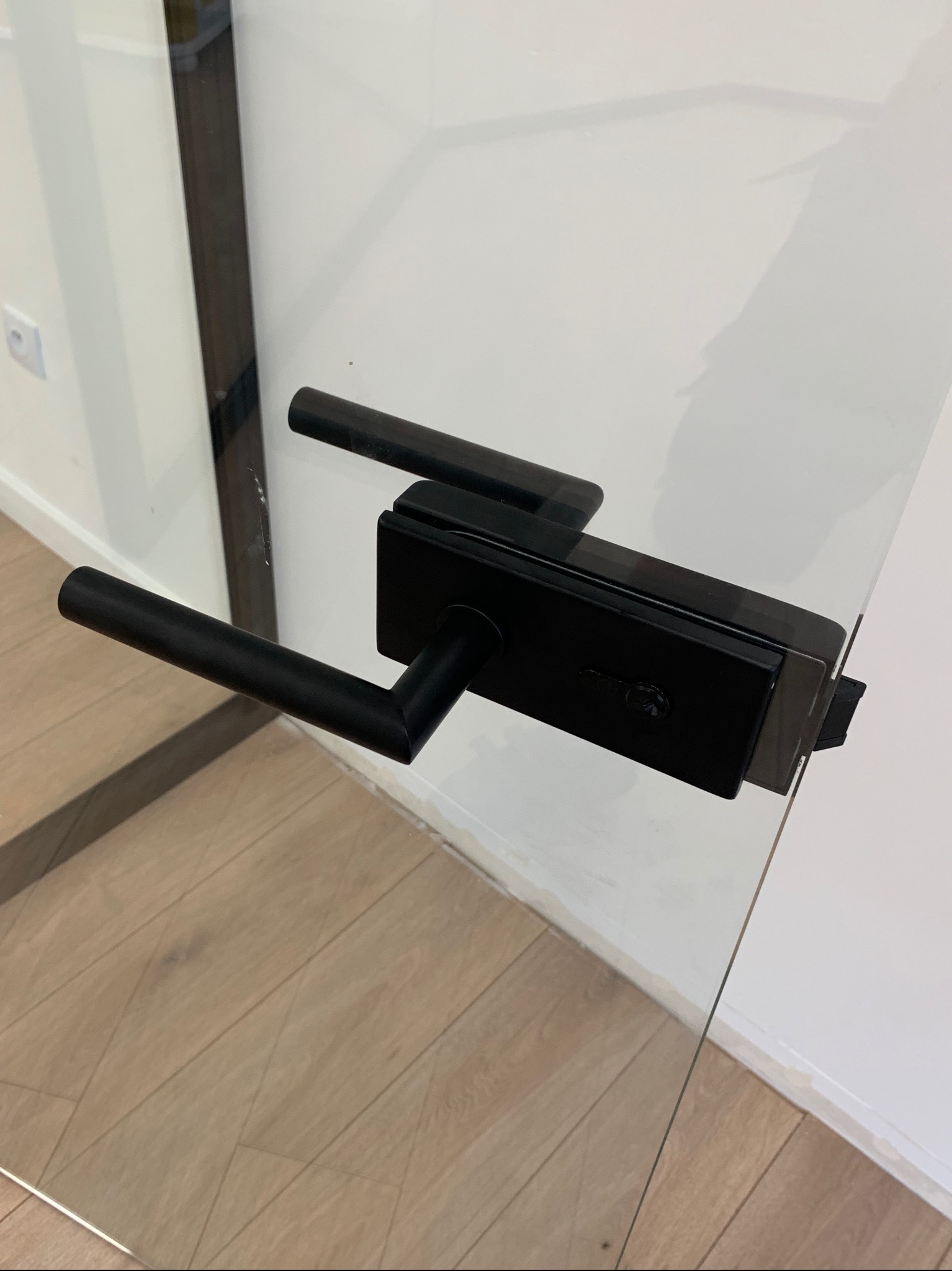 Szklane drzwi z czarną, nowoczesną klamką i zamkiem, widoczne na tle drewnianej podłogi i białej ściany.