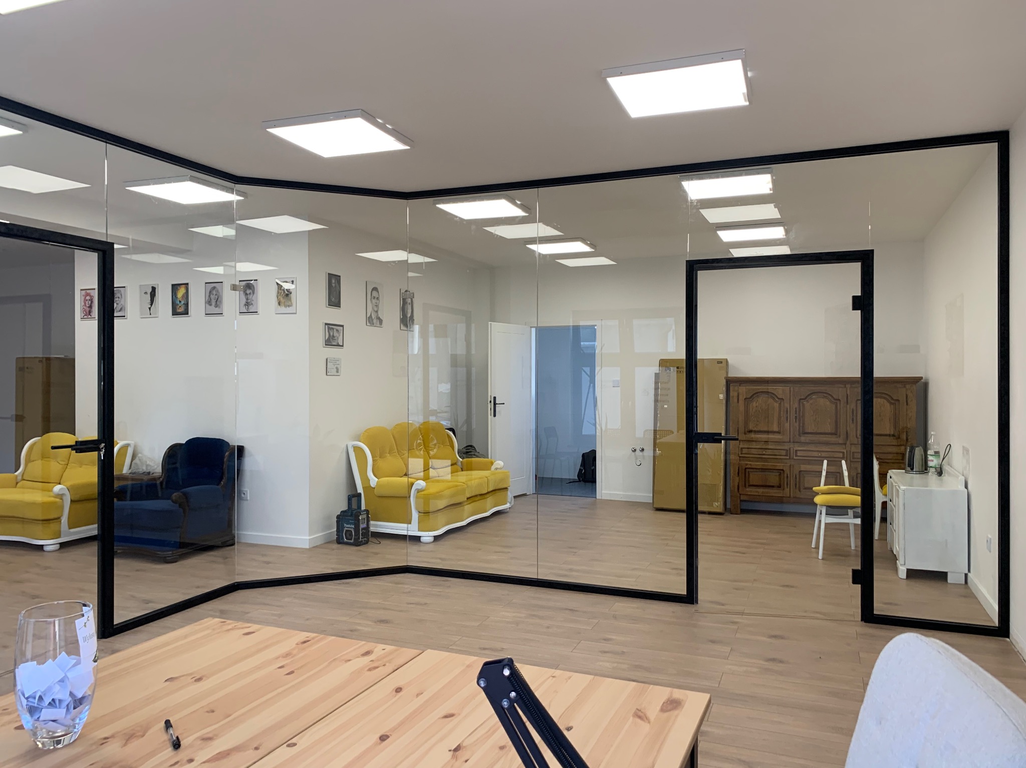 Nowoczesne biuro z przeszklonymi ścianami z czarnymi ramami, widoczne meble w stylu retro, drewniana podłoga i jasne oświetlenie.