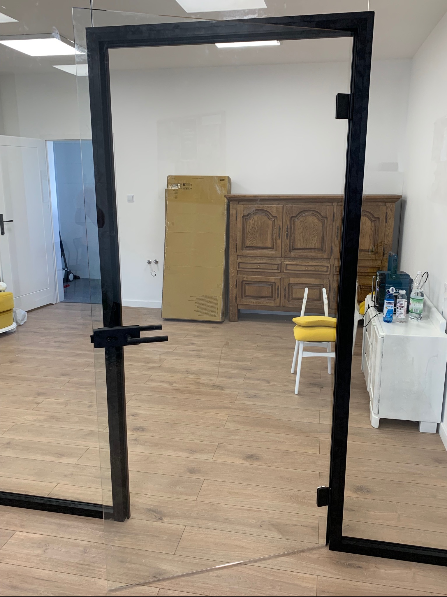 Szklane drzwi z czarną ramą w nowoczesnym wnętrzu, widoczne elementy wyposażenia: drewniana komoda, biała komoda, krzesło z żółtą poduszką i kartonowe pudło.