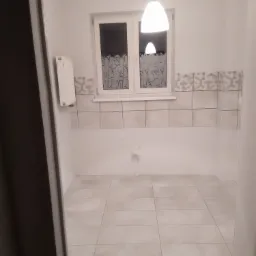 Płytki podłogowe i ścienne w jasnych odcieniach szarości w nowo wykończonym pomieszczeniu, widoczny grzejnik i okno z dekoracyjną folią, surowy otwór na kran, wisząca lampa.