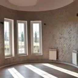 Wnętrze okrągłego pokoju z trzema wysokimi oknami, ściany pokryte dekoracyjnym tynkiem w odcieniach beżu i szarości, drewniana podłoga z widocznymi promieniami słońca, dwa białe grzejniki na ścianach.