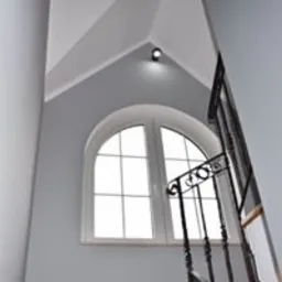 Wnętrze klatki schodowej z oknem łukowym i metalową balustradą, świeżo pomalowane na szaro, z widoczną lampą sufitową punktową.