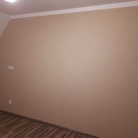 Beżowa ściana w pomieszczeniu po malowaniu, widoczne gniazdka elektryczne, biały pas dekoracyjny pod sufitem i fragment podłogi z panelami.