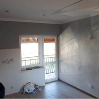 Pokój po częściowym malowaniu ścian na szaro, widoczne niedomalowane fragmenty, okno balkonowe z czarną barierką, na podłodze szare płytki, w rogu torba z materiałami budowlanymi.