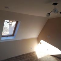 Poddasze po remoncie: skośna ściana pomalowana na beżowo, okno dachowe z widokiem na komin i błękitne niebo, drewnopodobna podłoga, widoczne ślady po szpachlowaniu na ścianie, lampa sufitowa w rogu.