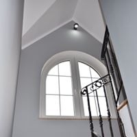 Wnętrze klatki schodowej z oknem łukowym i metalową balustradą, świeżo pomalowane na szaro, z widoczną lampą sufitową punktową.