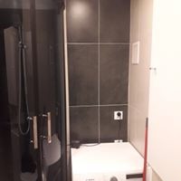 Ciemnoszara łazienka z kabiną prysznicową po lewej stronie i pralką pod ciemnymi płytkami ściennymi. Widoczny biały kontakt nad pralką. Minimalistyczny styl.