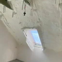 Poddasze po ociepleniu pianką PUR, widoczne krokwie, metalowe uchwyty i okno dachowe zabezpieczone folią