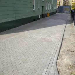 Nowa, szara kostka brukowa ułożona wzdłuż budynku z zieloną elewacją i metalowym ogrodzeniem, widok z perspektywy chodnika.