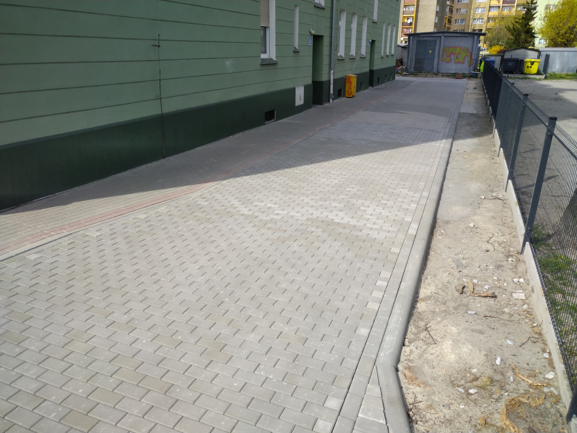 Nowa, szara kostka brukowa ułożona wzdłuż budynku z zieloną elewacją i metalowym ogrodzeniem, widok z perspektywy chodnika.