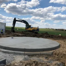 Okrągła, świeżo wylana płyta fundamentowa z widocznymi prętami zbrojeniowymi, na tle krajobrazu z błękitnym niebem i chmurami, z koparką Volvo w tle.