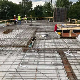 Siatka zbrojeniowa na betonowej płycie fundamentowej z instalacją elektryczną, pracownicy w odblaskowych kamizelkach konsultujący projekt w tle.