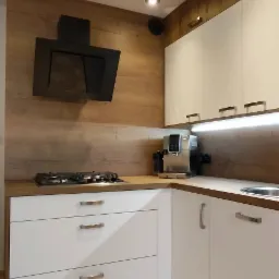 Nowoczesna kuchnia z białymi frontami, drewnianym blatem i okładziną ścienną, czarnym okapem i zabudowaną płytą gazową, oświetleniem podszafkowym i beżową podłogą z płytek.