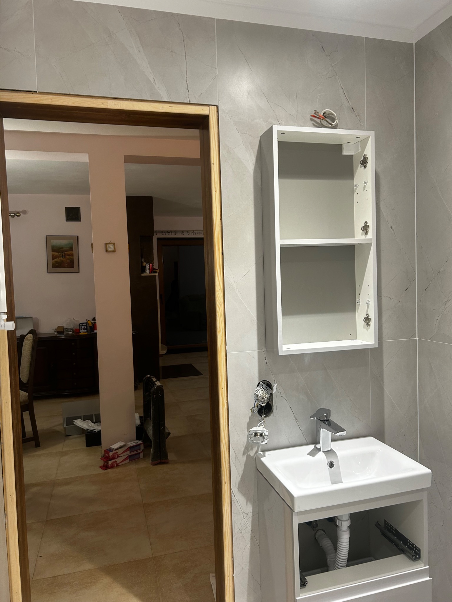 Łazienka w trakcie wykańczania: zamontowana szafka z umywalką, wisząca szafka bez drzwi, widoczne przewody elektryczne, ściany wyłożone szarymi płytkami imitującymi marmur, widok z korytarza.