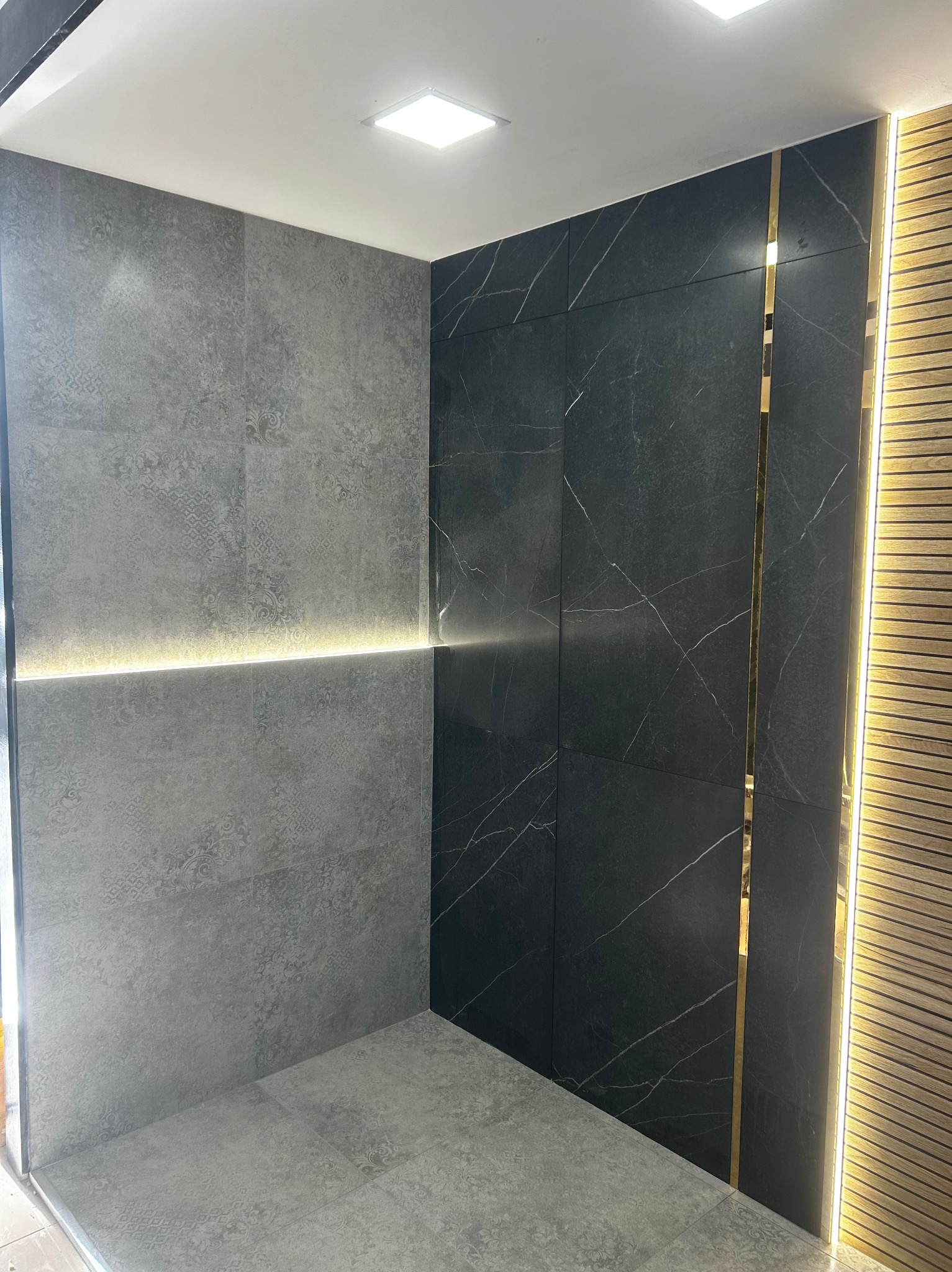 Nowoczesna łazienka z szarymi płytkami z subtelnym wzorem, czarnymi płytkami imitującymi marmur z złotymi paskami dekoracyjnymi oraz oświetleniem LED wzdłuż ściany z drewnianymi lamelami, tworząc...