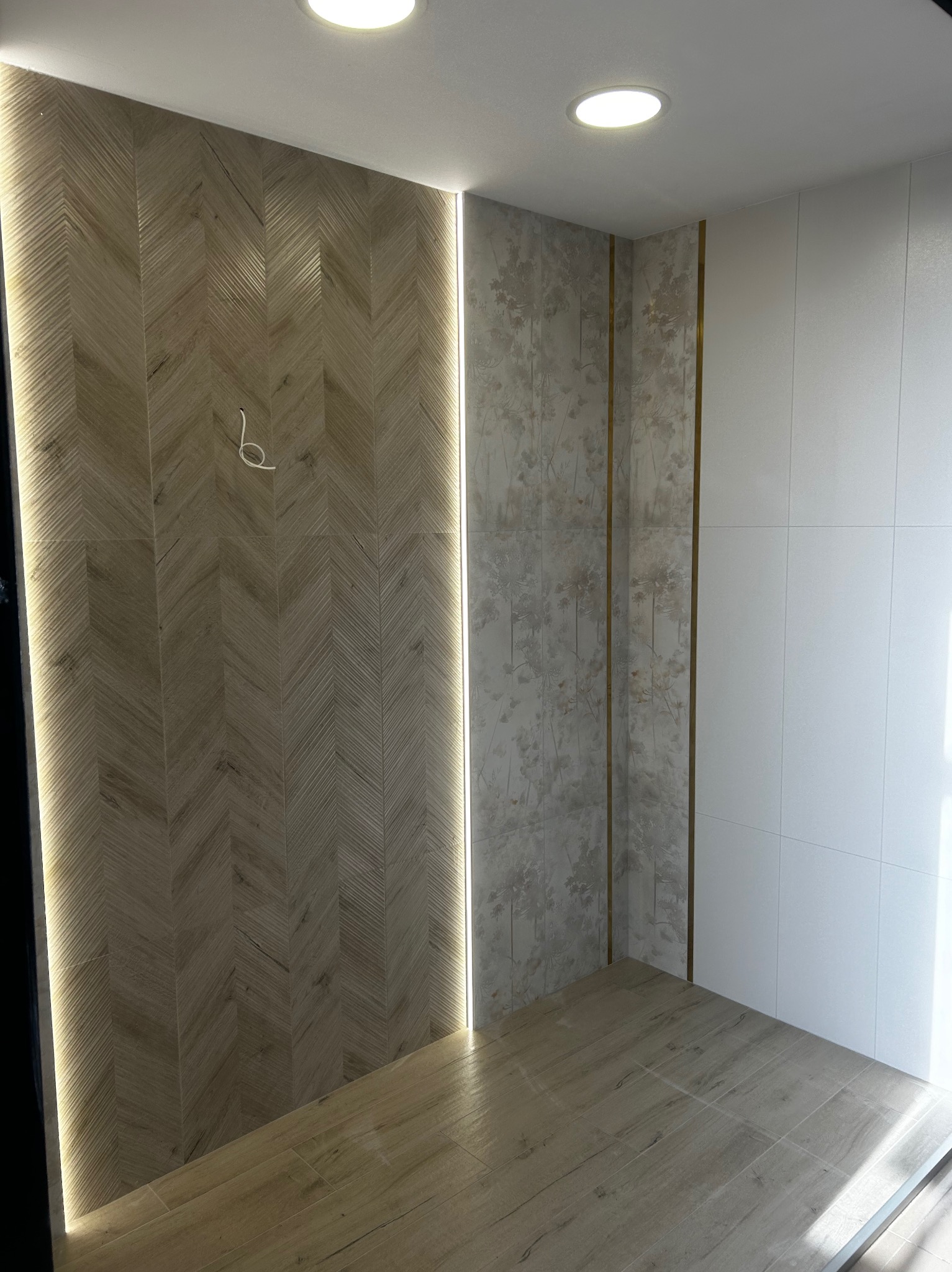 Wykończona łazienka z podświetlaną ścianą z płytek ułożonych w jodełkę, naprzeciwko ściana z białych płytek i ściana z tapetą w motywy roślinne z delikatnymi złotymi akcentami.