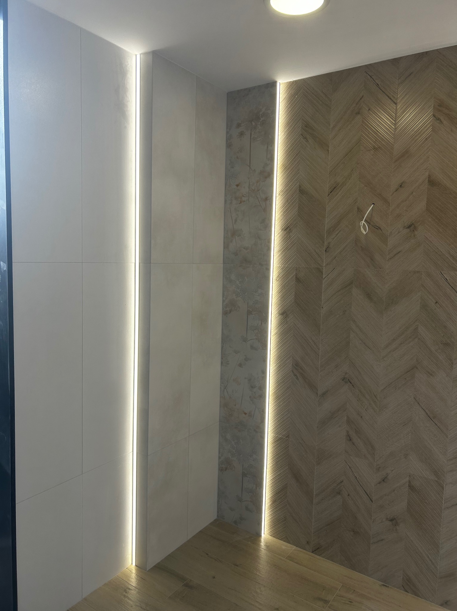 Nowoczesna łazienka z podświetlanymi panelami ściennymi, białe płytki kwadratowe, wzorzysta tapeta z motywem roślinnym i drewnopodobne płytki ułożone w jodełkę, widoczne przewody elektryczne