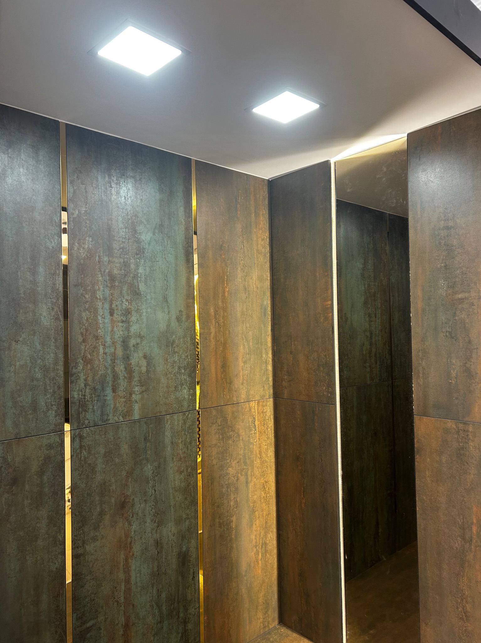 Rustykalne, wielkoformatowe płytki ścienne z metalicznym połyskiem, ułożone pionowo z wąskimi, złotymi listwami dekoracyjnymi, oświetlone dwoma kwadratowymi lampami sufitowymi.