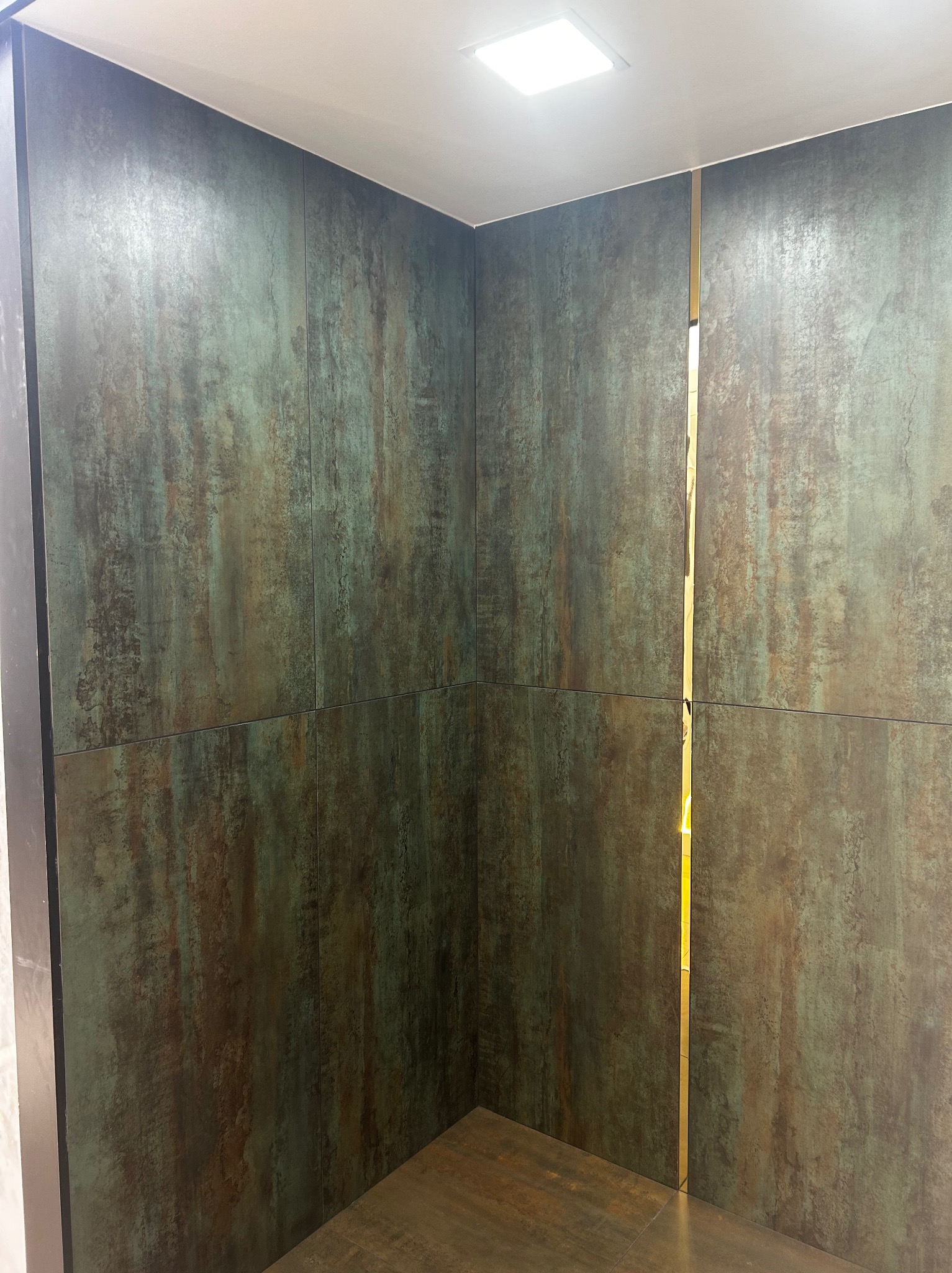 Ściana wyłożona dużymi płytami imitującymi rdzewiejący metal, z wąską, pionową, złotą listwą dekoracyjną między płytami, oświetlona górnym światłem.