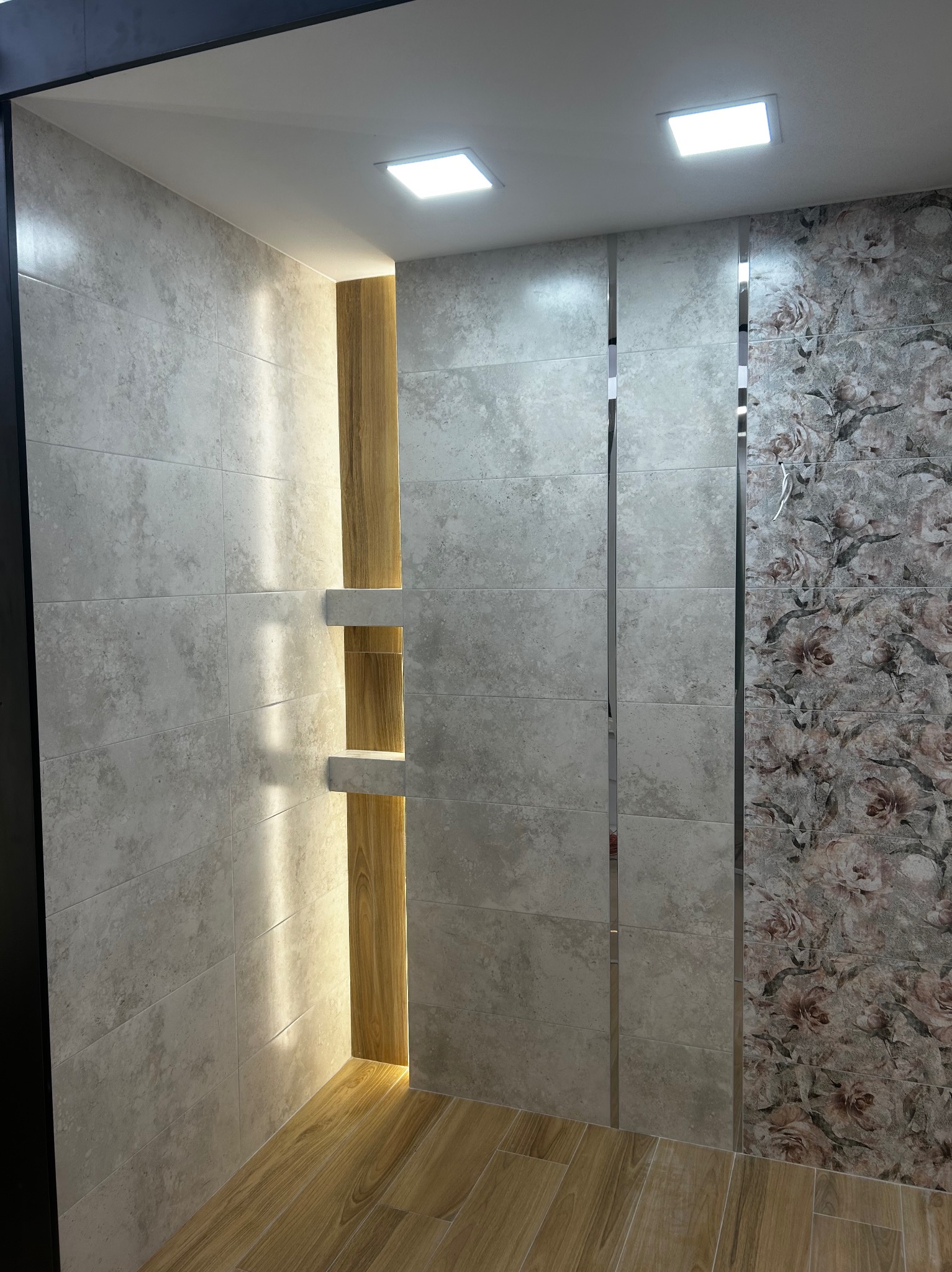 Wystawa płytek ceramicznych w łazience z oświetloną niszą z drewnianym akcentem i podłogą z paneli drewnopodobnych, prezentująca różne wzory i tekstury.