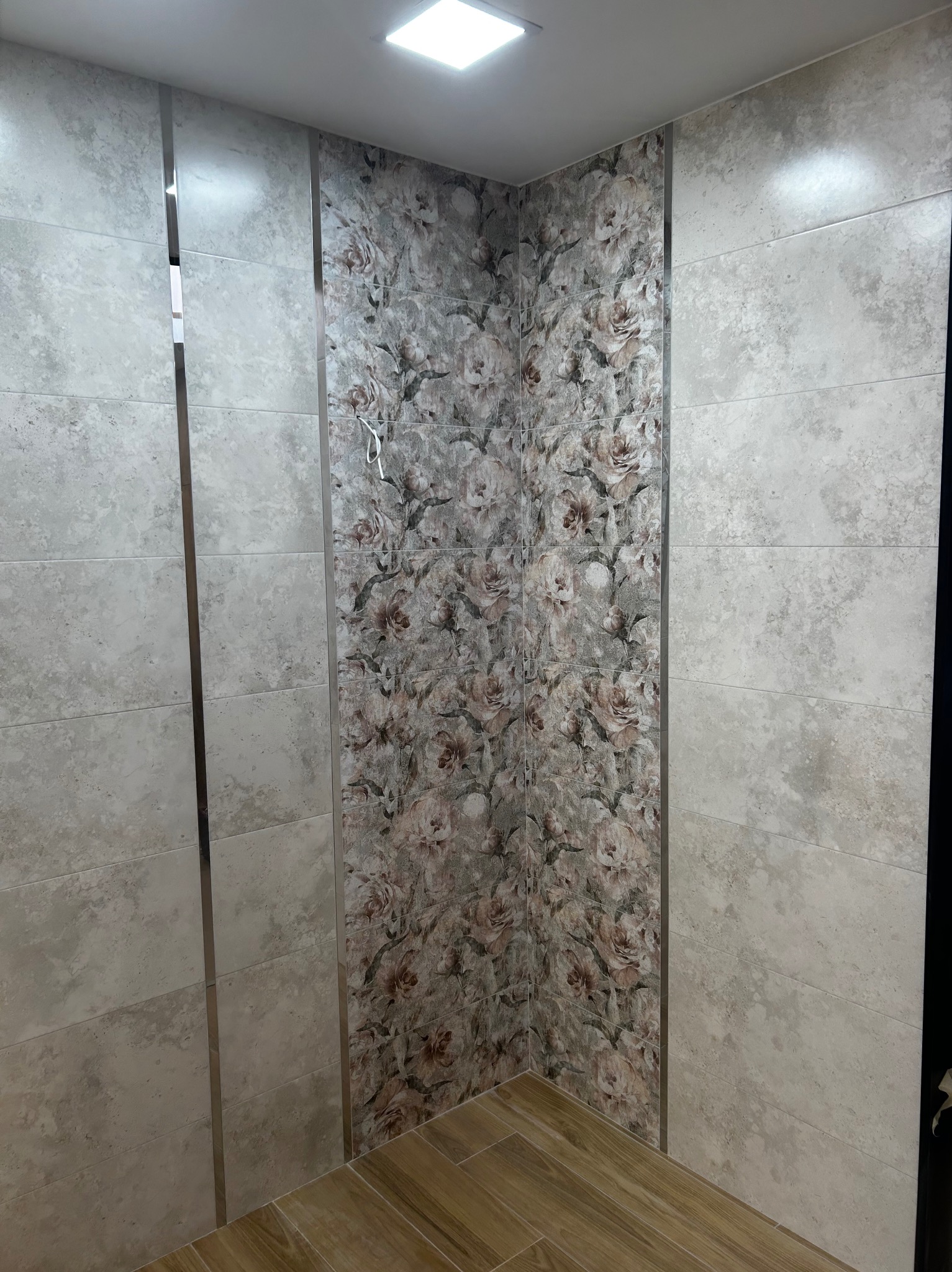 Wykończony narożnik łazienki z płytkami w odcieniach beżu i szarości, z dekoracyjnym pasem płytek w kwiatowy wzór, oddzielonych metalowymi listwami, oraz drewnopodobną podłogą.