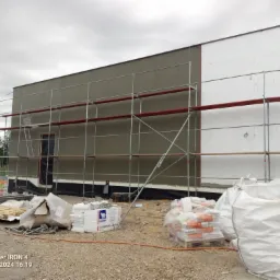 Budynek w trakcie ocieplania elewacji: rusztowanie, częściowo ocieplona ściana styropianem, worki z materiałami budowlanymi i paczki styropianu na żwirowym podłożu.