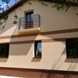 Dwukondygnacyjny dom z elewacją w odcieniach beżu i brązu, z balkonem z metalową balustradą, widok przez gałęzie drzew