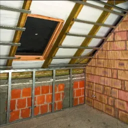 Wnętrze poddasza w Kłobucku w trakcie prac: widoczne krokwie z membraną, okno dachowe w drewnianej ramie oraz metalowy stelaż do montażu płyt gipsowo-kartonowych na ścianie z częściowo odsłoniętą...