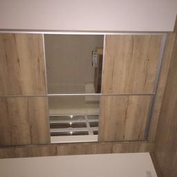 Kemoiz pracownia stolarska - Wnętrze szafy przesuwnej z drewnopodobnymi drzwiami, widok na okno przez otwarte drzwi szafy.