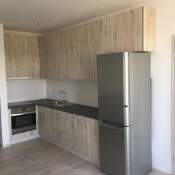 Kemoiz pracownia stolarska - Jasna kuchnia na wymiar z drewnianymi frontami i szarym blatem, widoczna lodówka ze stali nierdzewnej, jasna podłoga i drzwi z delikatnym wzorem drewna.