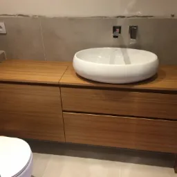 Biała, owalna umywalka nablatowa na szafce z drewnopodobną okleiną, minimalistyczna bateria umywalkowa w kolorze chromu, szare płytki na ścianie, widoczny fragment toalety.