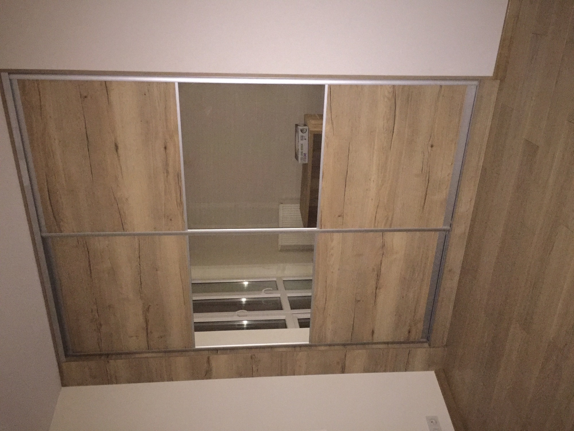 Wnętrze szafy przesuwnej z drewnopodobnymi drzwiami, widok na okno przez otwarte drzwi szafy.