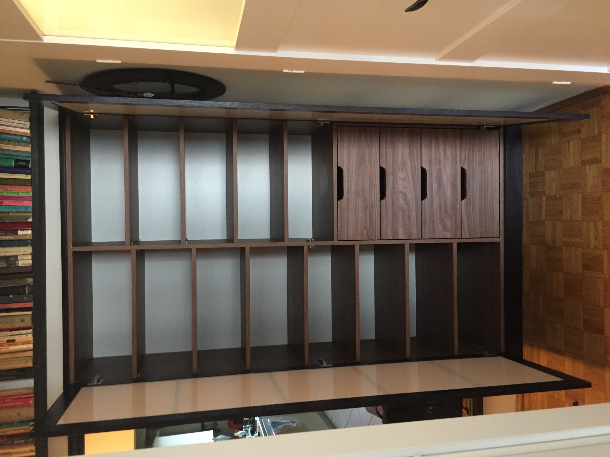 Otwarta szafa z licznymi półkami i trzema drewnianymi drzwiczkami, widoczna z dołu, obok regał z książkami i parkiet w jodełkę.