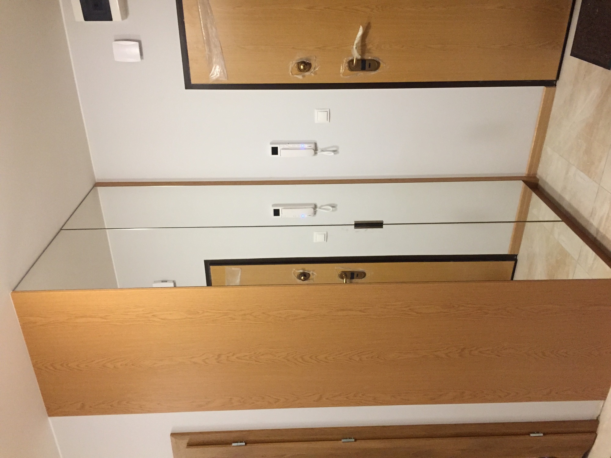 Wnętrze przedpokoju z lustrem na ścianie, odbijającym fragment drewnianych drzwi z czarną ramą i interkomem na białej ścianie.