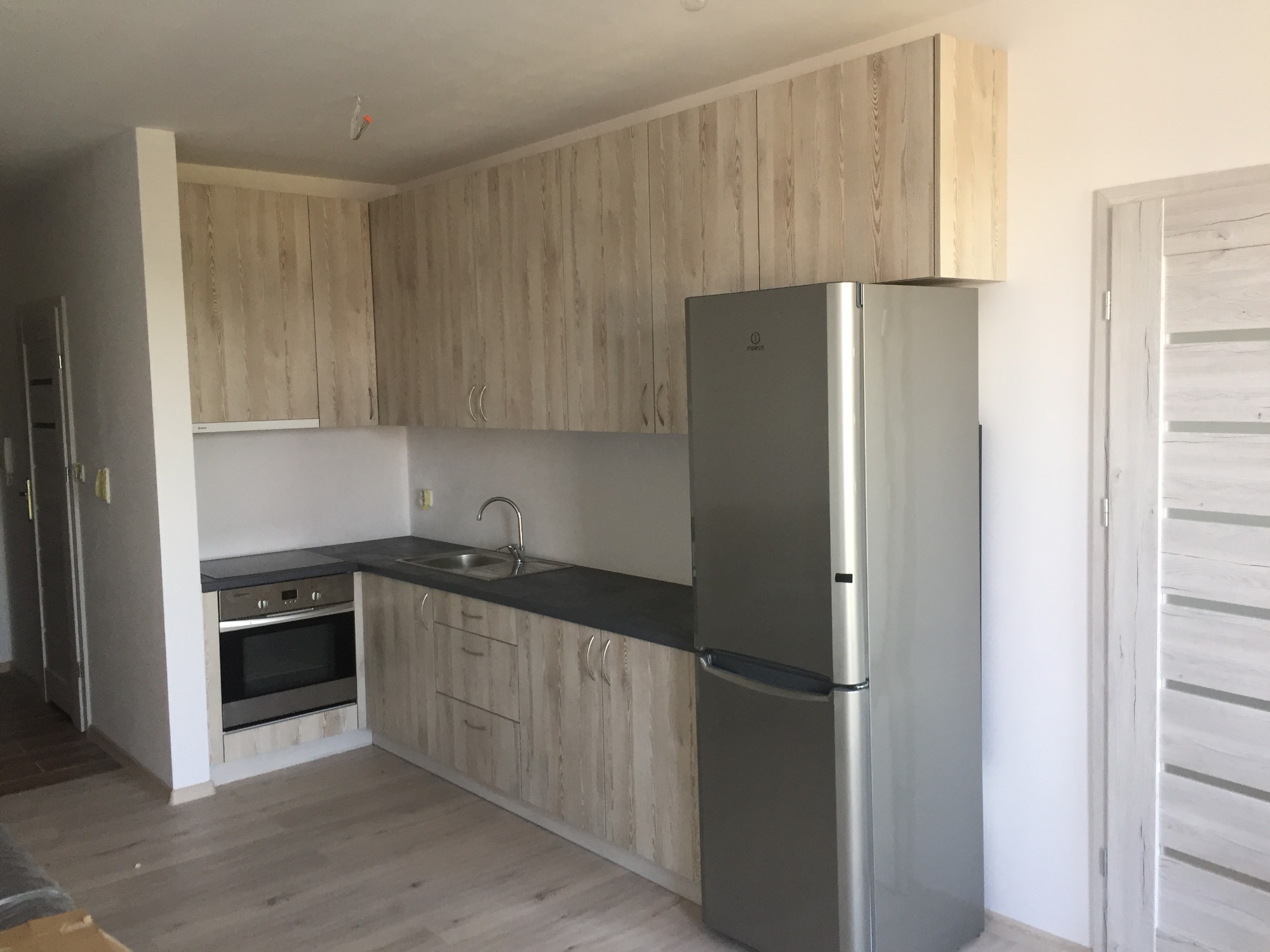 Jasna kuchnia na wymiar z drewnianymi frontami i szarym blatem, widoczna lodówka ze stali nierdzewnej, jasna podłoga i drzwi z delikatnym wzorem drewna.