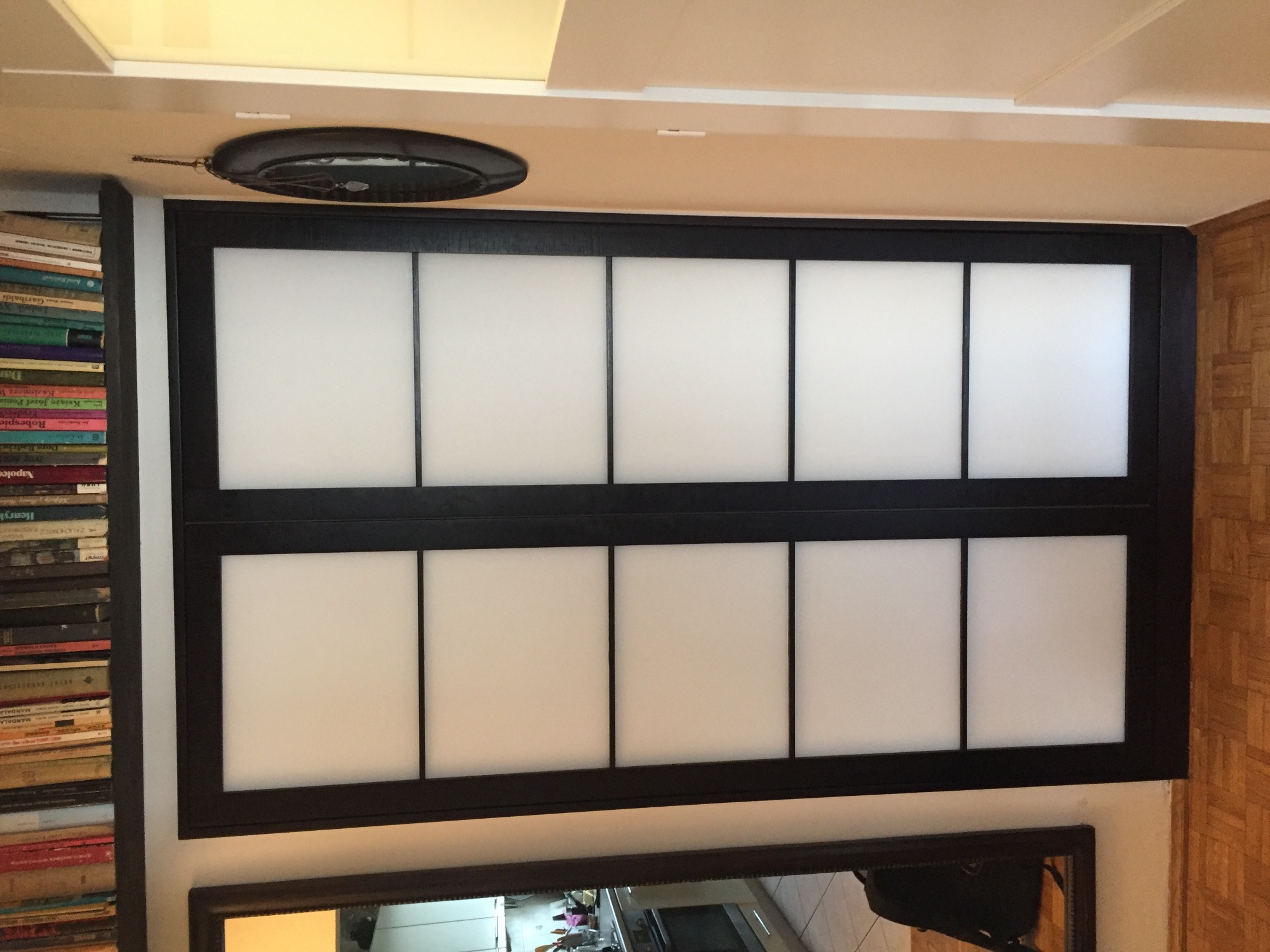 Przesuwne drzwi do szafy z czarną ramą i białymi panelami, widoczne książki na półce obok i odbicie kuchni w lustrze poniżej.